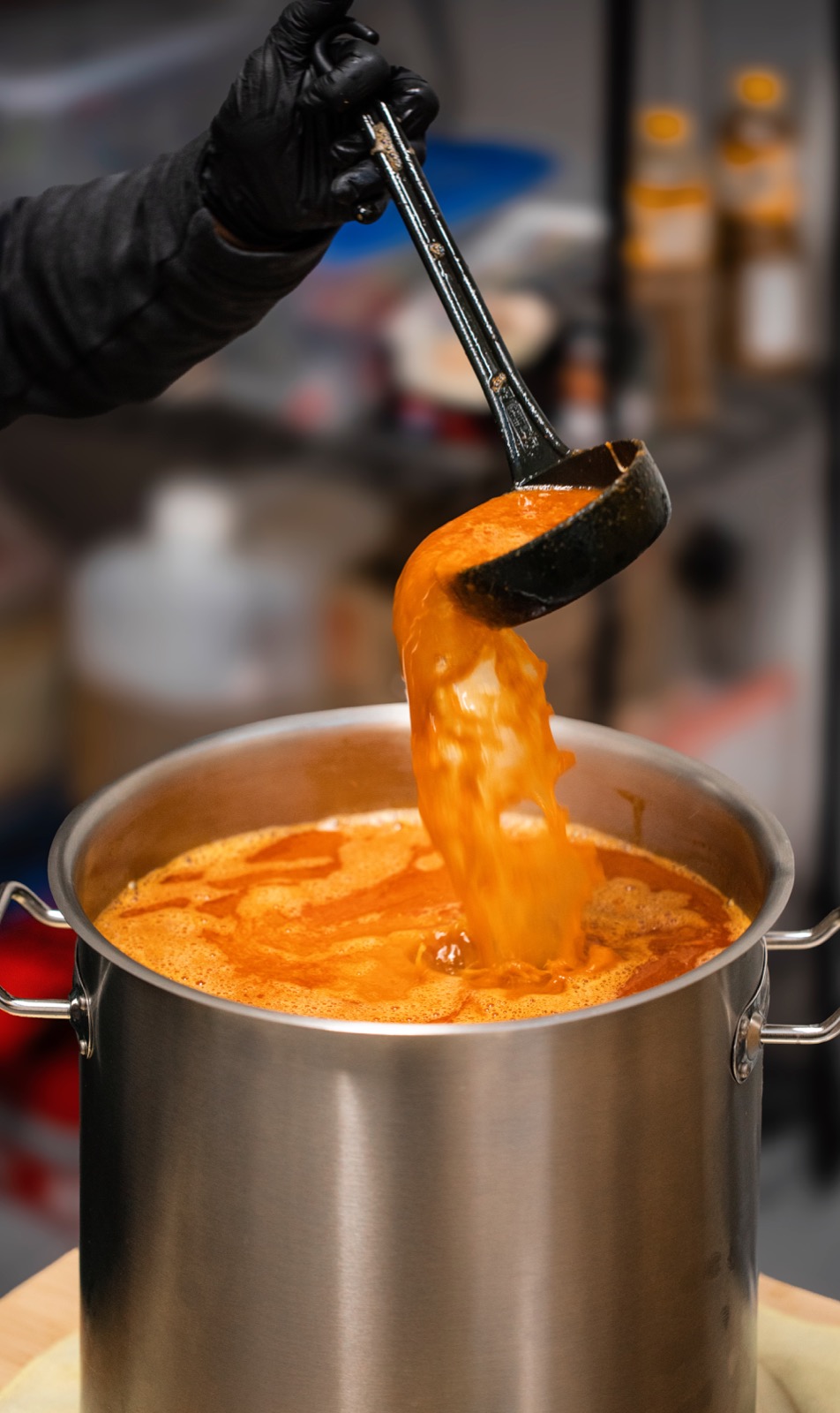 A chef in black gloves ladling rich orange broth into a large steel pot.